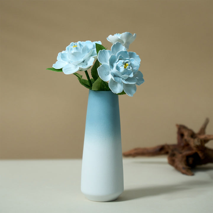 Four pale blue porcelain peonies in a sky-blue-to-white ombre vase on a white surface against a beige wall.