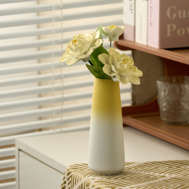 Porcelain flower set with three pale yellow peonies in a yellow-to-cream tapered ceramic vase by a sunlit window.