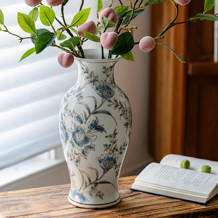 Tall vase with blue floral motifs, thin gold-colored outlines, and pink fruiting branches on a wooden table by a window.
