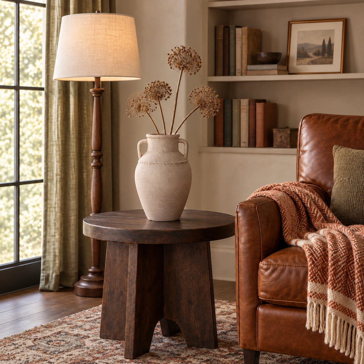 Textured beige vase with dry seed stems on a dark oak table beside a tobacco leather reading chair