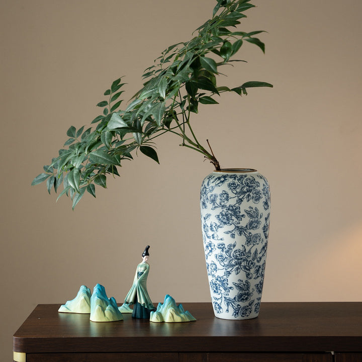 Tall blue and white floral vase holding leafy green branches, displayed on a dark wood console beside ornaments.