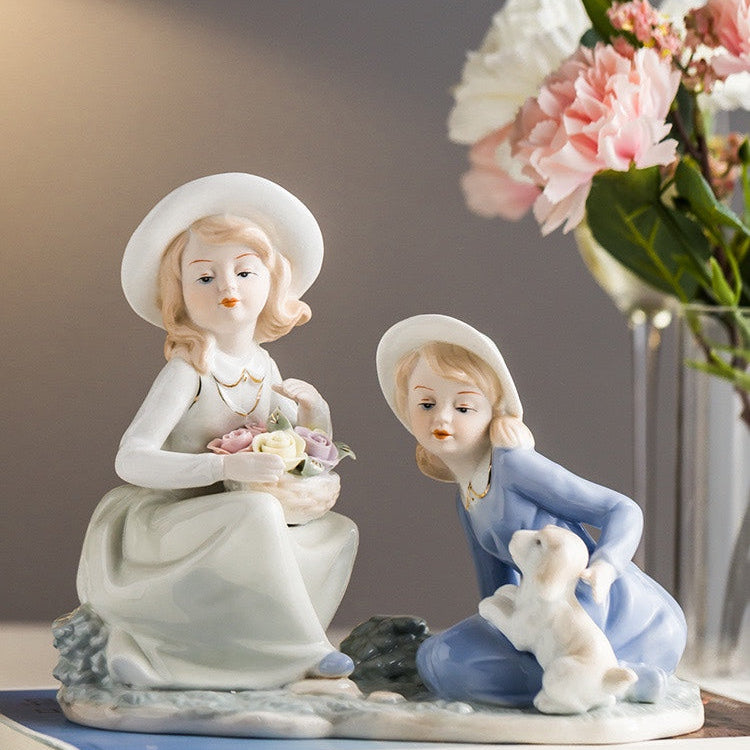 Two ceramic figurines of children with flowers and a teddy bear on a book, with flowers in the background.