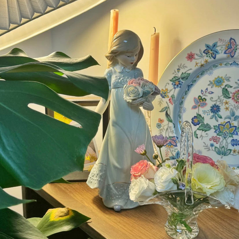 Decorative figurine of a woman holding flowers on a table with a floral plate and plant.