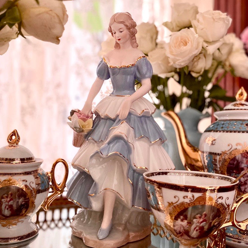 Decorative tea set with a figurine of a woman in a blue dress on a table with flowers.