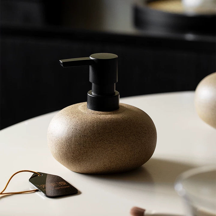 Beige speckled ceramic pebble soap dispenser with a matte black pump, standing alone on a cream tabletop.