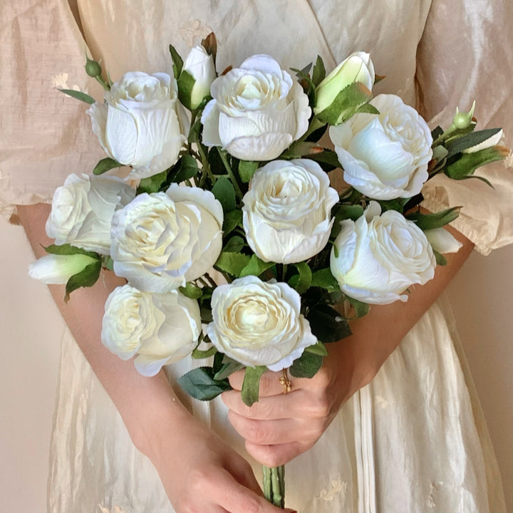 Full bouquet of white faux roses with open layered blooms and upright buds held against a cream dress.