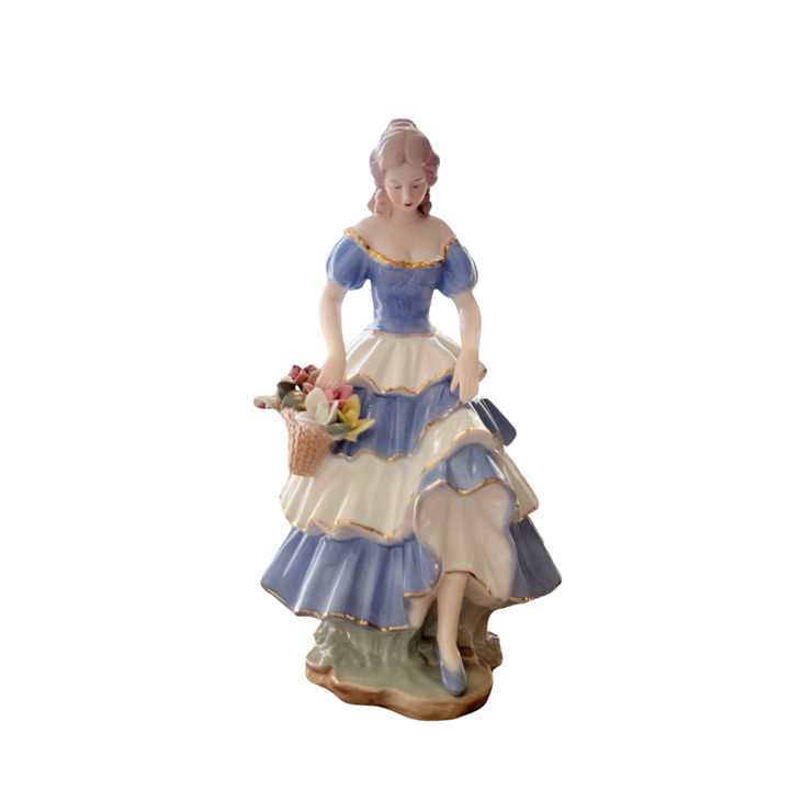 Seated porcelain lady figurine in a blue-and-white tiered dress with gold trim and flower basket on a white background.