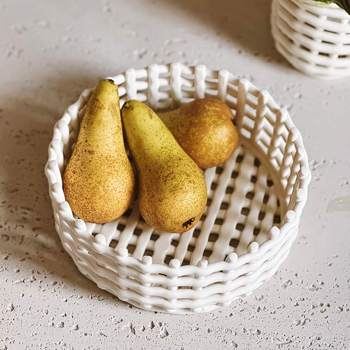 High-angle view of a white round ceramic lattice basket holding three golden-green pears on a countertop.