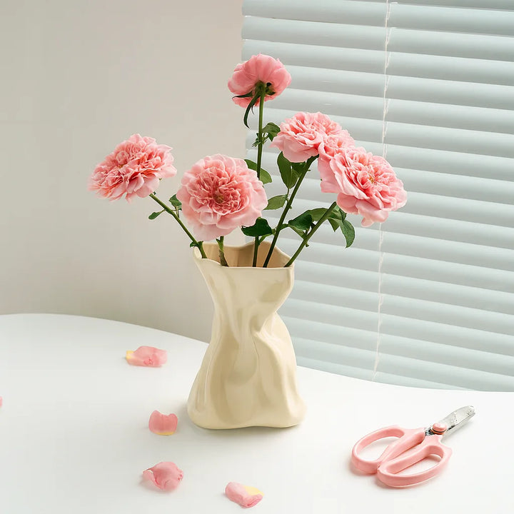 Cream crinkled ceramic vase holding five pink roses on a white table before sunlit window blinds.
