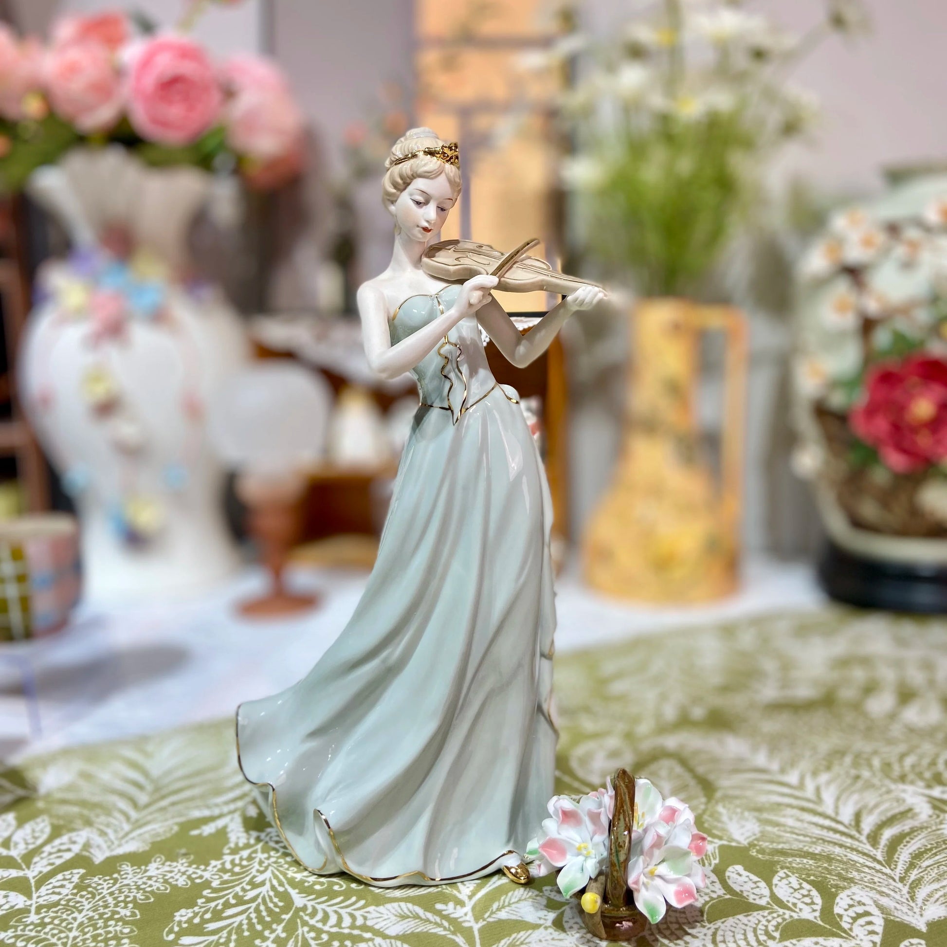 Decorative figurine of a woman holding a violin on a table with floral arrangements in the background.