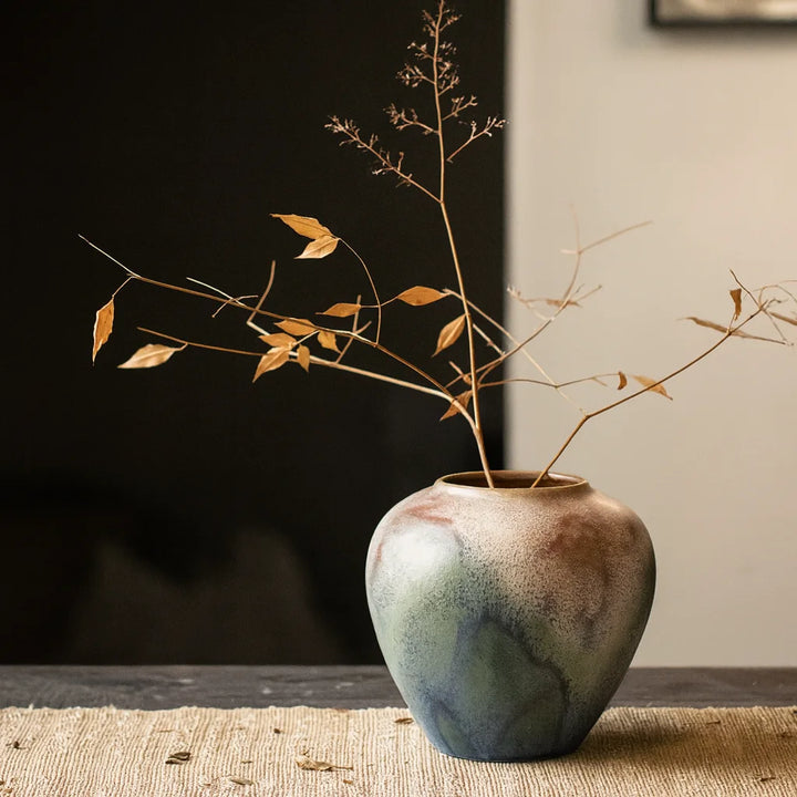 Round reactive-glaze ceramic vase in cream, blue, and green with dried stems on a woven runner