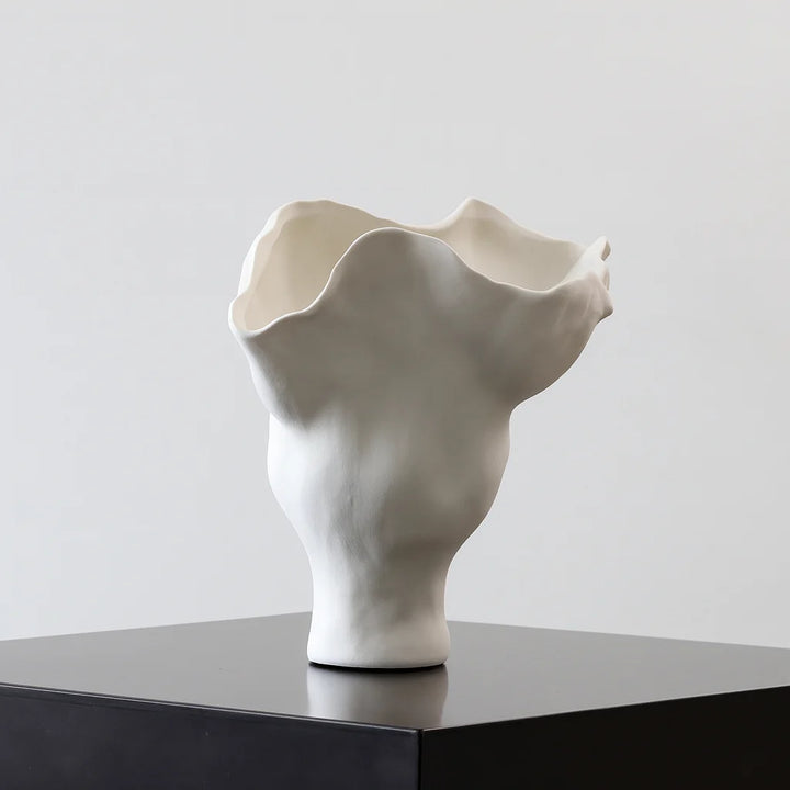 Short off-white sculptural ruffle vase with an undulating folded rim, displayed empty on a black plinth.