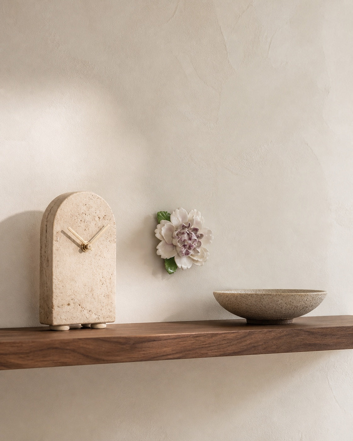One small plum-purple Ruffled Peony ceramic wall flower beside a travertine clock and low bowl on a walnut shelf.