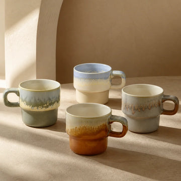 Four ceramic mugs in green, golden, rust and pale-blue glazes arranged on a linen-covered table.