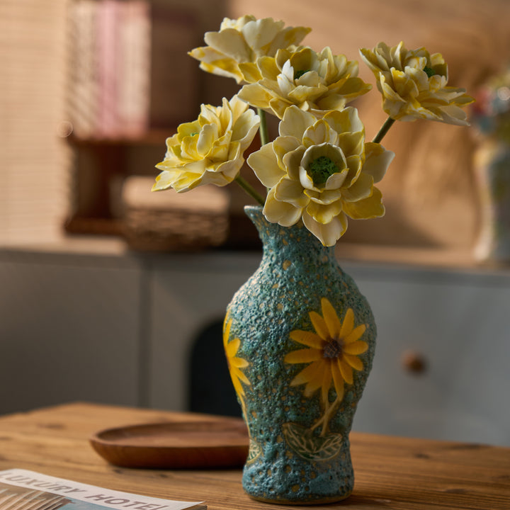 Six cream-and-yellow peony diffuser flowers in a turquoise floral ceramic vase on a sunlit wood table.