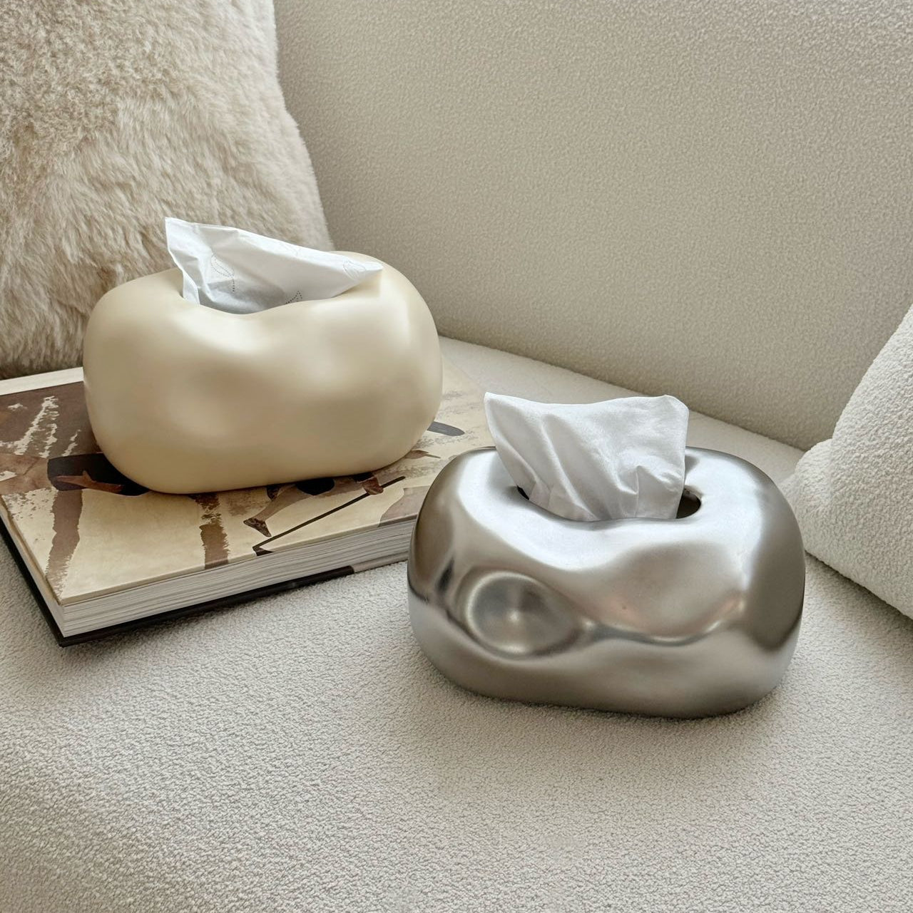 Two tissue boxes, one white and one silver, on a couch with a book underneath.