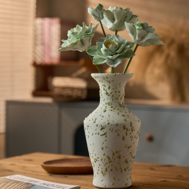 Five pale mint ceramic flowers in a cream vase with moss-green speckles, arranged on a wood table in a warm interior.