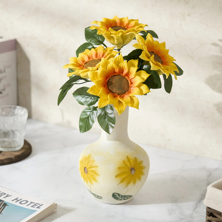 Porcelain sunflower bouquet of four yellow blooms in a painted cream vase, staged on a tabletop beside a glass.