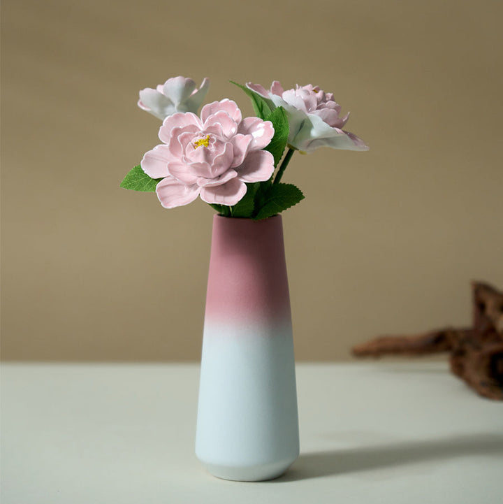 Pink ceramic peony bouquet with green leaves in a pink-to-white gradient vase on a pale tabletop