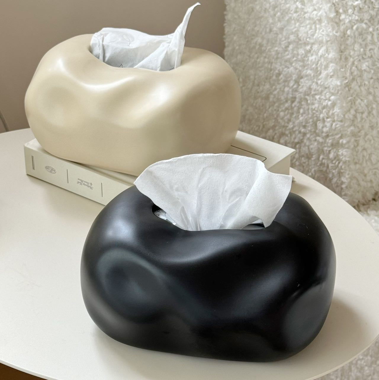 Two unique tissue holders, one beige and one black, on a table with tissues inside.