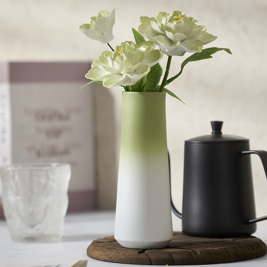 Gradient green and white vase with flowers on a table next to a black teapot.