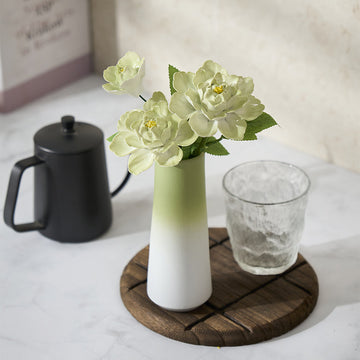 White vase with green accents holding flowers on a wooden coaster with a glass and black container in the background.