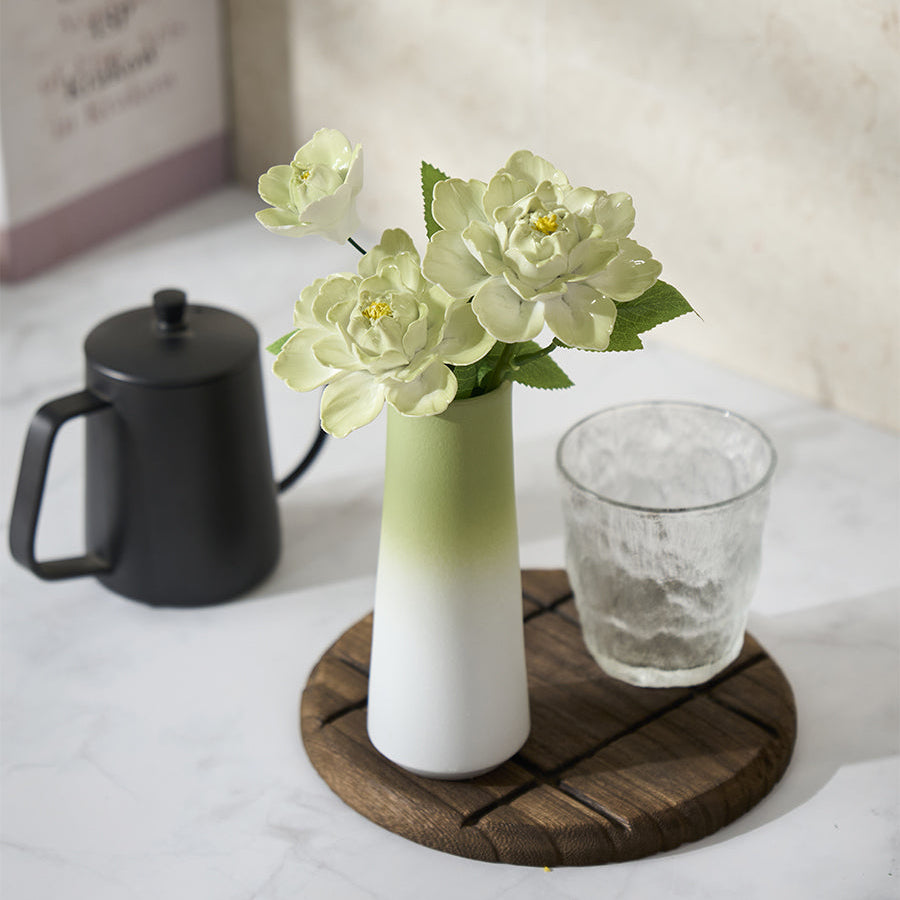 White vase with green accents holding flowers on a wooden coaster with a glass and black container in the background.