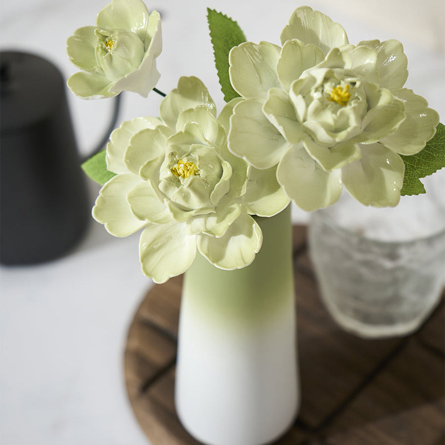 White vase with green accents holding flowers on a wooden coaster with a glass and black container in the background.
