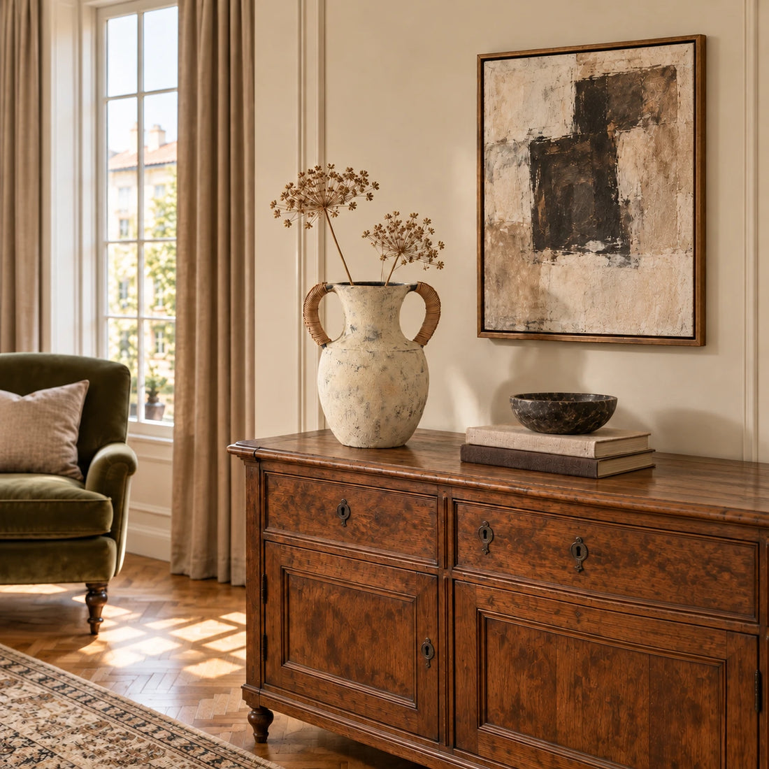 Weathered twin-handle ceramic vase with dried stems on a walnut cabinet beside an olive armchair