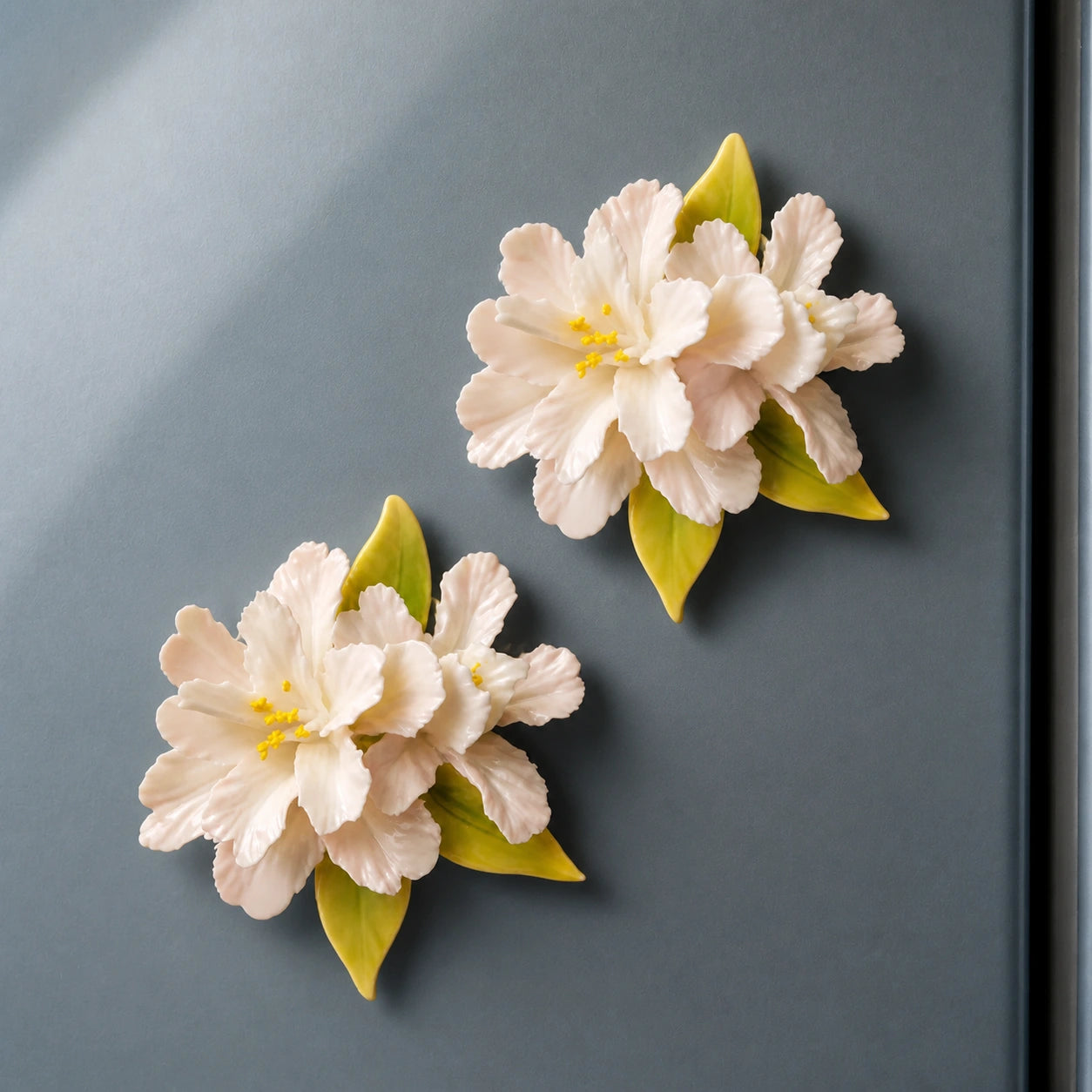 Two blush-white ceramic crabapple magnets arranged as a collector pair on a mineral blue-gray refrigerator.