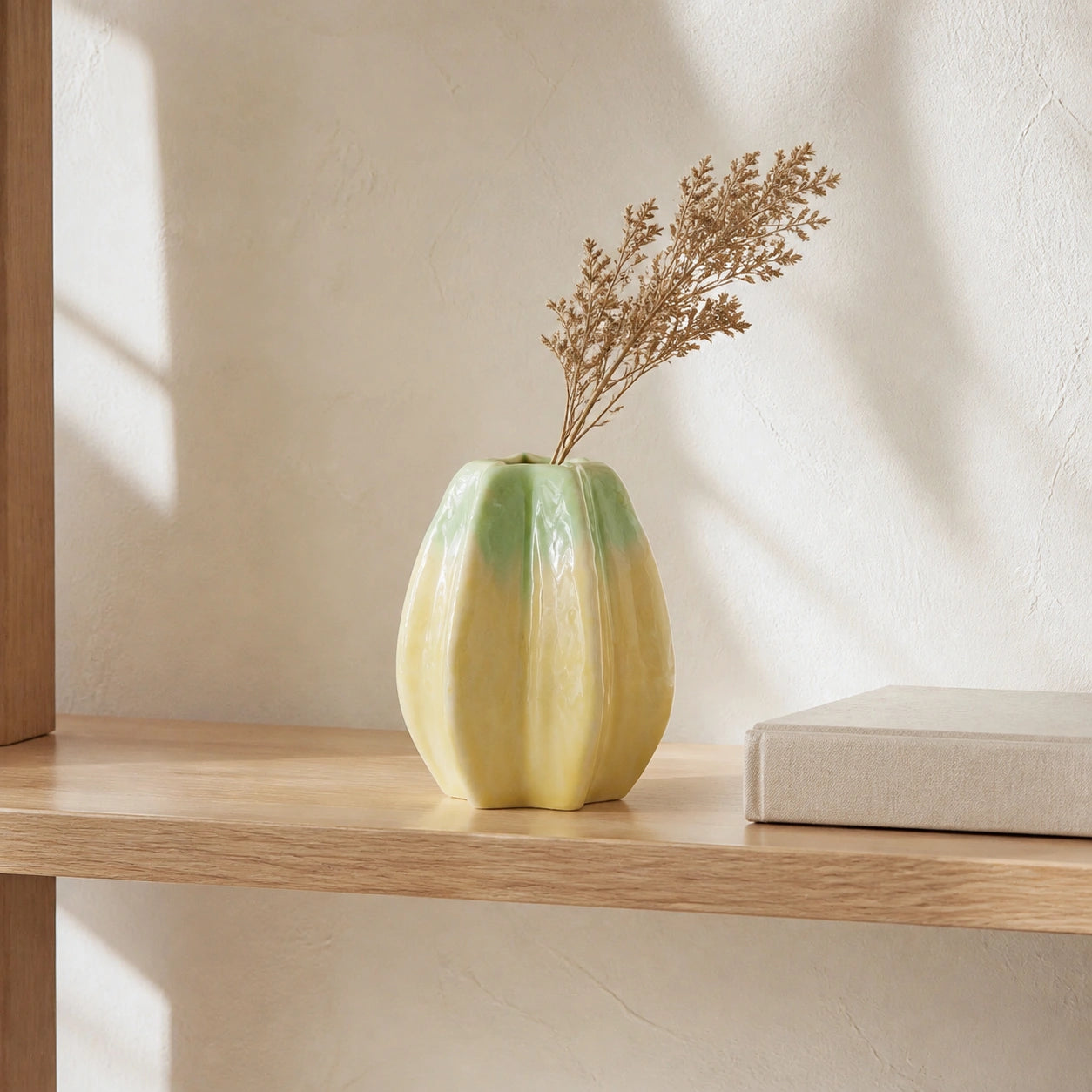 Small yellow-green lobed ceramic vase styled with a dried stem on a pale oak shelf.