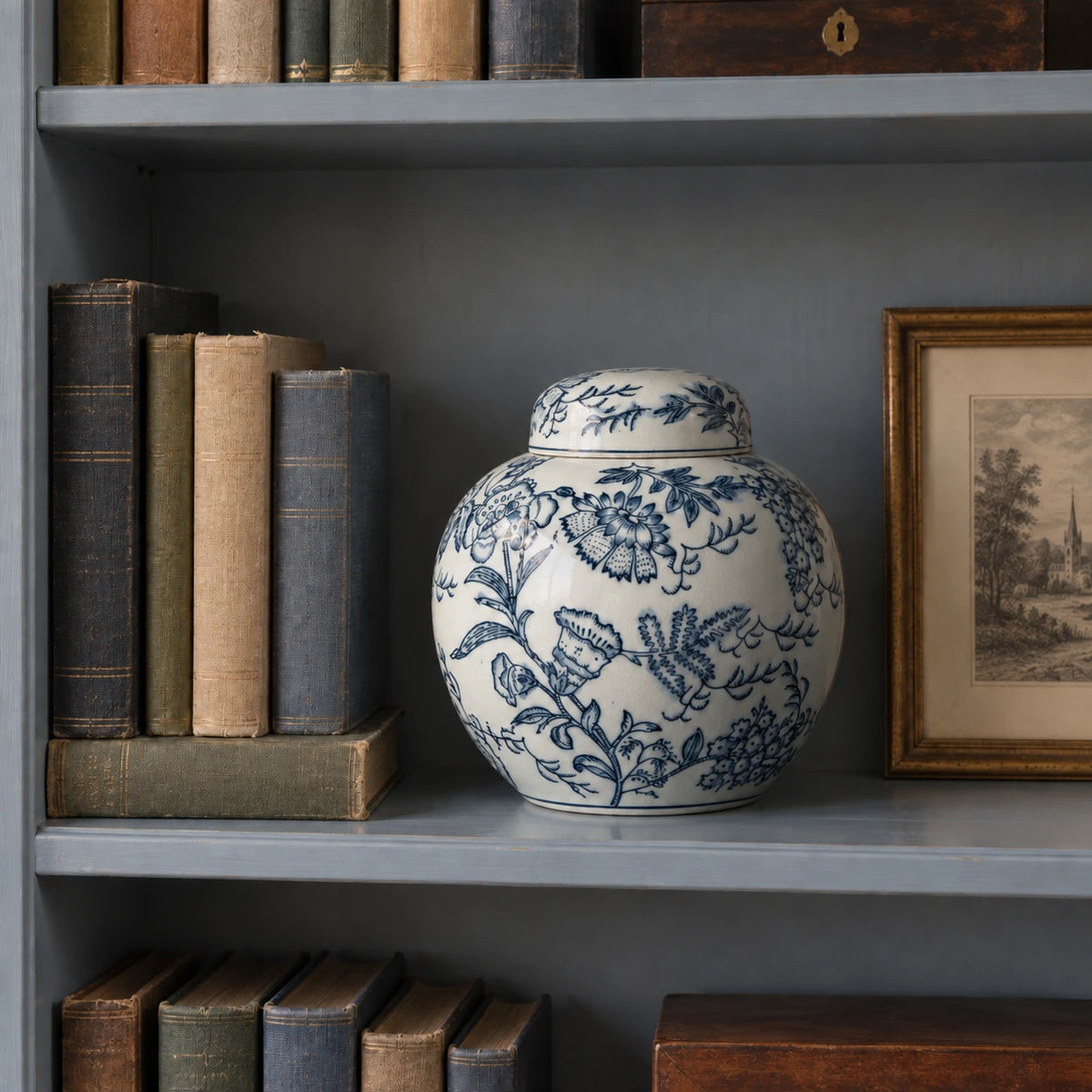 Geschlossenes blau-weißes Blumengefäß auf einem blaugrauen Bücherregal mit alten Büchern und Radierung.