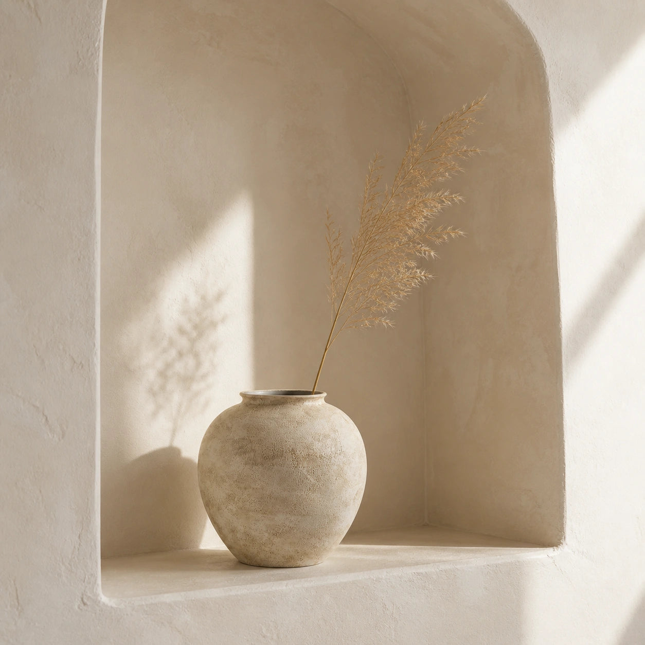 Small stone-textured ivory ceramic vase with one dried grass stem in a sunlit plaster niche.