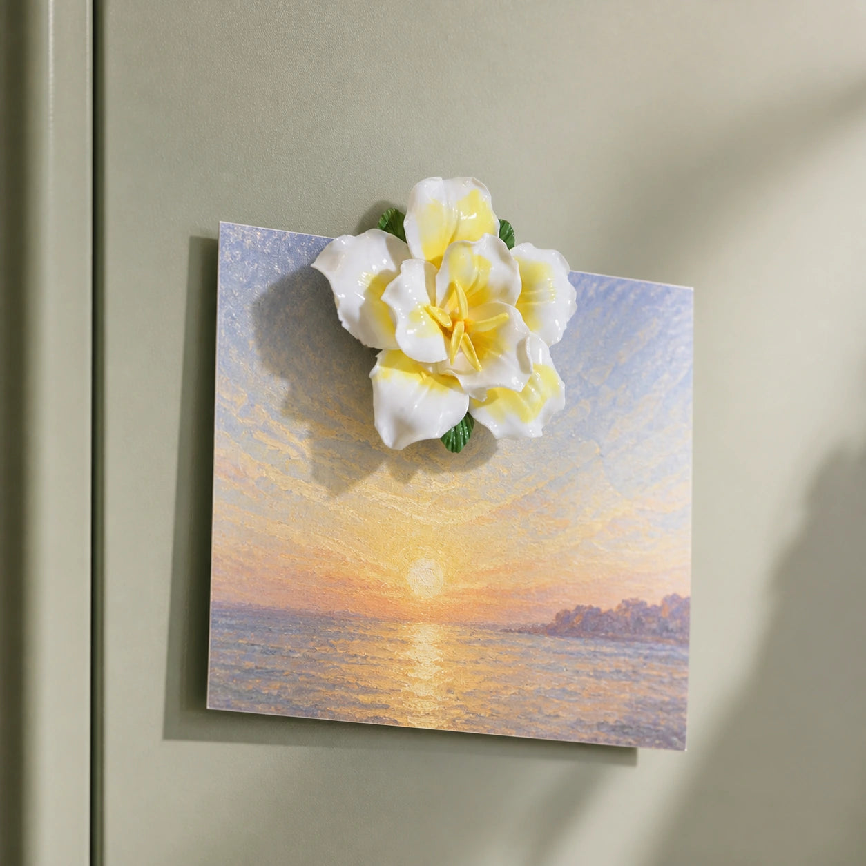Butter yellow ceramic lily magnet holding a painted sunrise card on a pale sage pantry door.