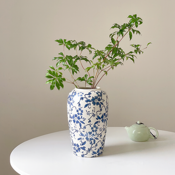 Tall white ceramic vase with blue floral patterns holding green leafy branches on a white round table next to a green teapot