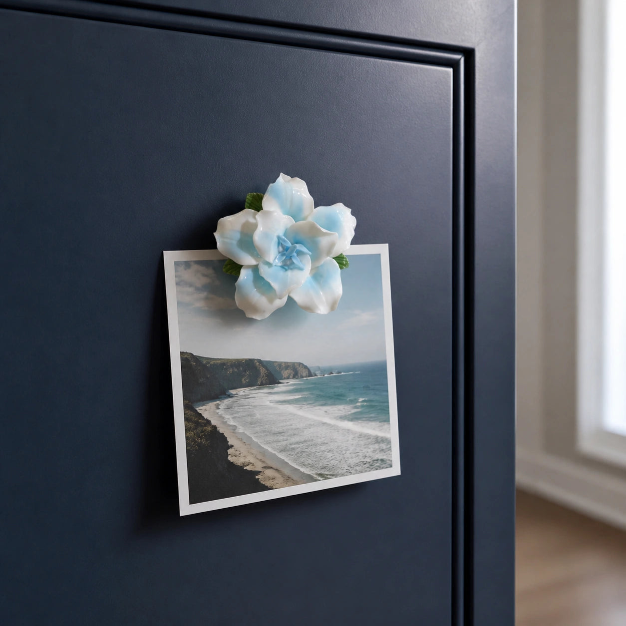 Powder blue ceramic lily magnet holding a coastal photograph on a deep navy cabinet.
