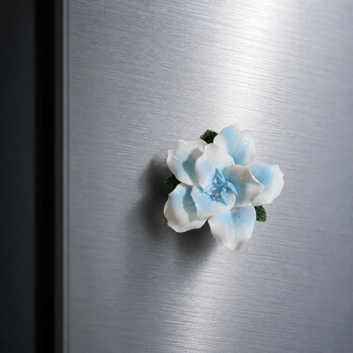 Powder blue ceramic lily magnet styled as a cool accent on brushed stainless steel.