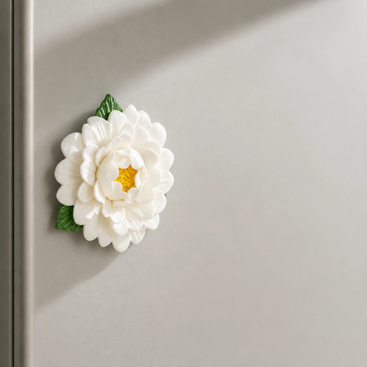 Aimant pivoine en céramique blanc chaud projetant une ombre sculpturale sur un réfrigérateur grège.