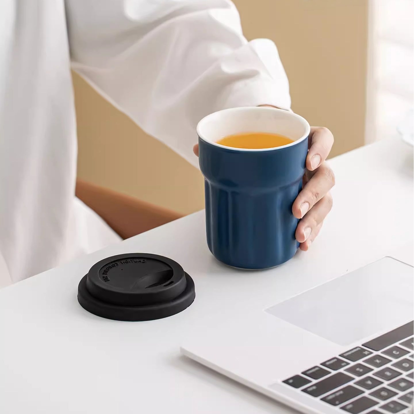 Navy blue fluted ceramic mug filled with tea beside black travel lid and laptop