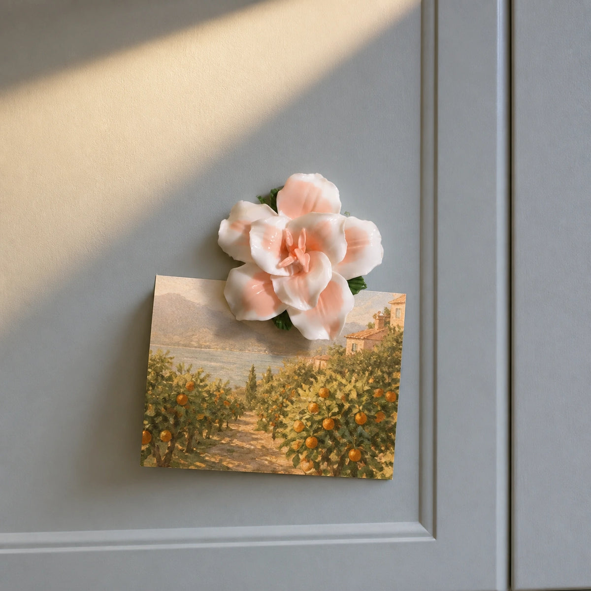 Soft orange ceramic lily magnet holding a warm orchard photograph on a blue-gray cabinet.