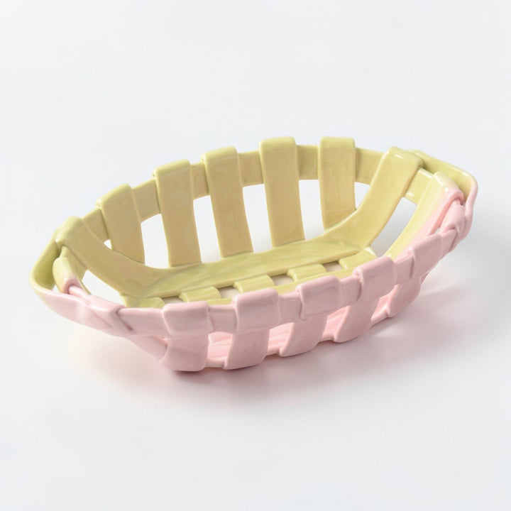 Shallow oval woven ceramic basket in pale pink and yellow shown empty from a front three-quarter angle.
