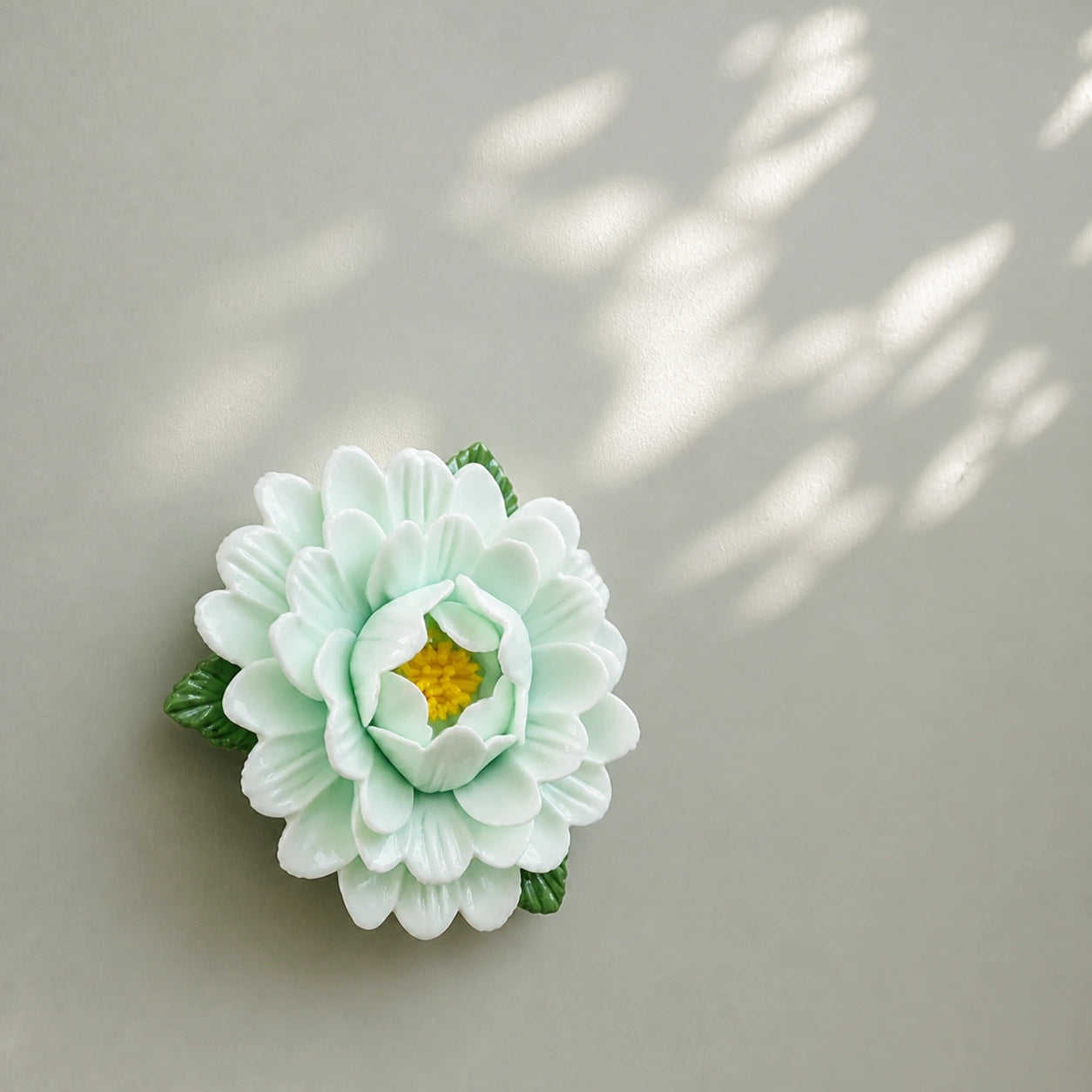 Aimant pivoine en céramique vert pâle sous de douces ombres de feuilles sur une surface gris sauge.