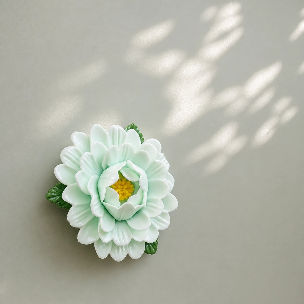Aimant pivoine en céramique vert pâle sous de douces ombres de feuilles sur une surface gris sauge.
