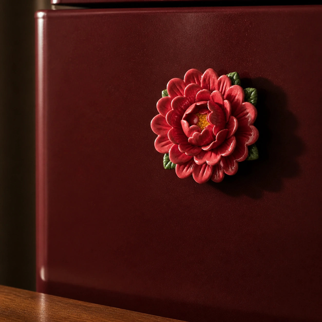 Aimant pivoine en céramique rouge rosé sur un mini-réfrigérateur bordeaux dans une lumière latérale chaude.