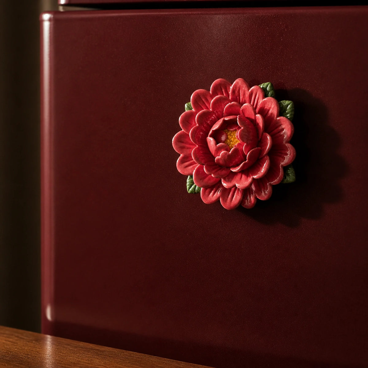 Aimant pivoine en céramique rouge rosé sur un mini-réfrigérateur bordeaux dans une lumière latérale chaude.