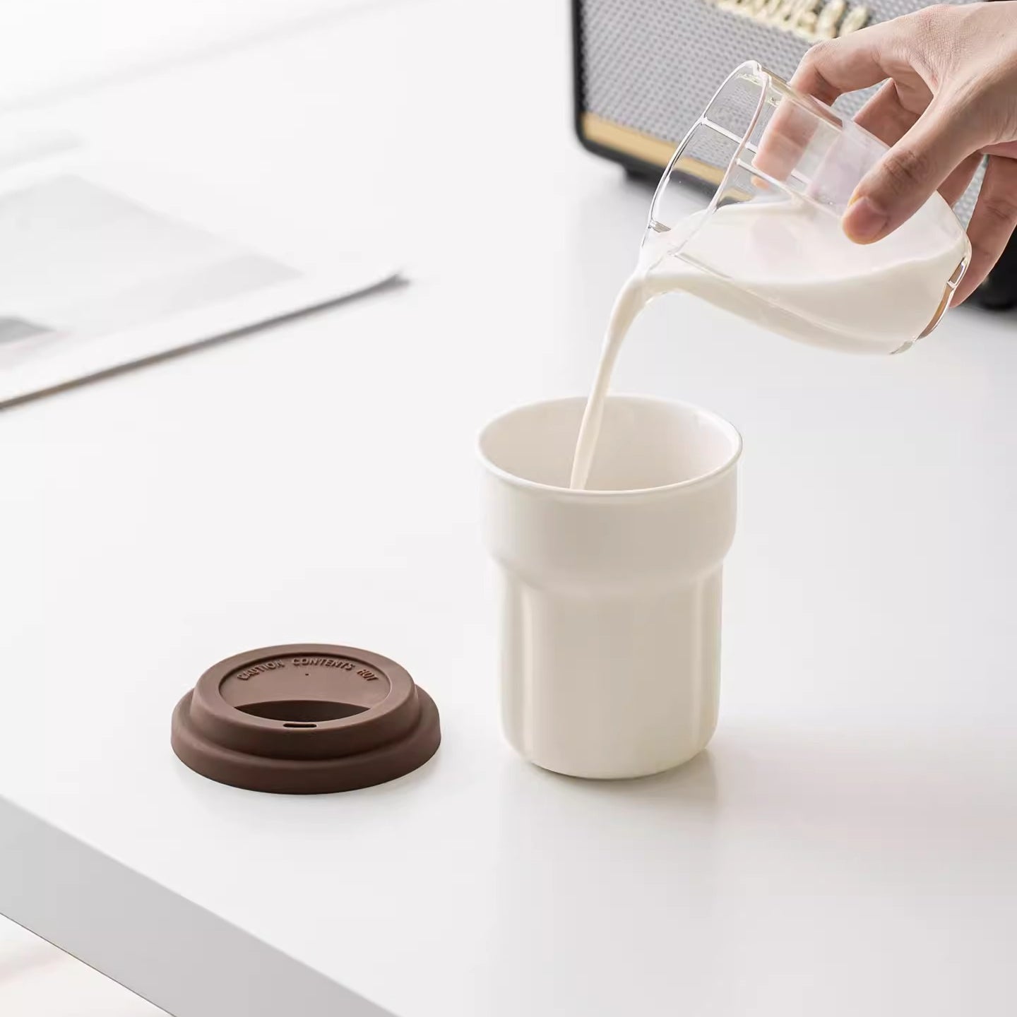 Milk being poured into a cream fluted ceramic mug beside its brown travel lid