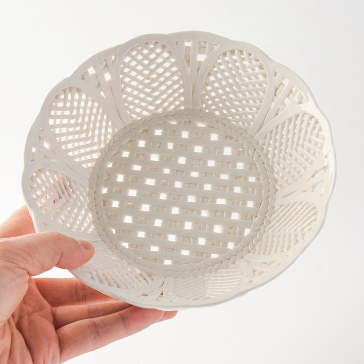 Hand holding an empty white ceramic lattice fruit bowl, showing repeating herringbone leaf panels and a scalloped rim.