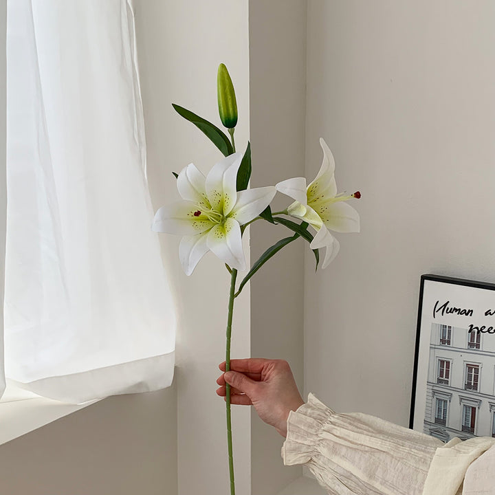 Hand holding a single artificial lily stem with two open white blooms, a green bud, and dark leaves.