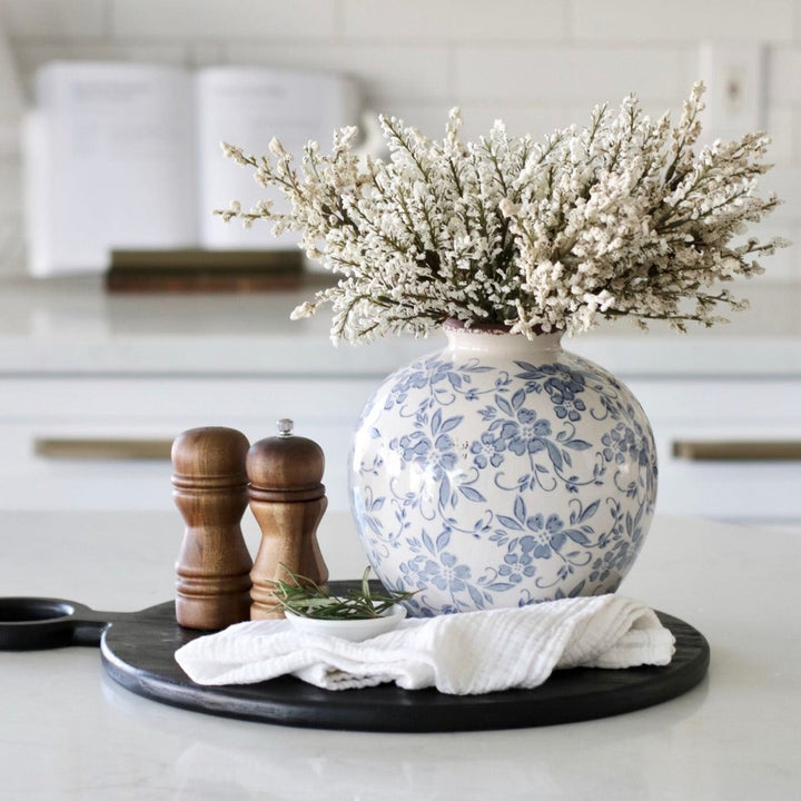 Round blue-and-white floral vase holding white blossoms on a black tray beside two wooden grinders in a sunlit kitchen.