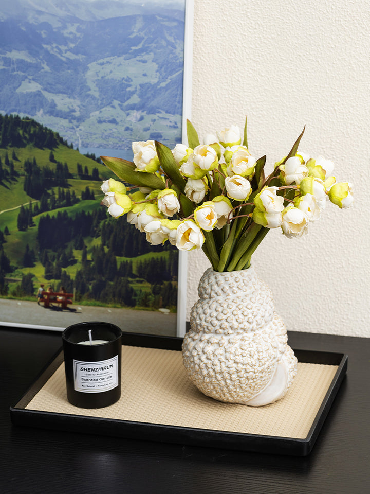 White conch-shaped ceramic vase holding white flower buds on a beige tray atop a black table beside a black candle.